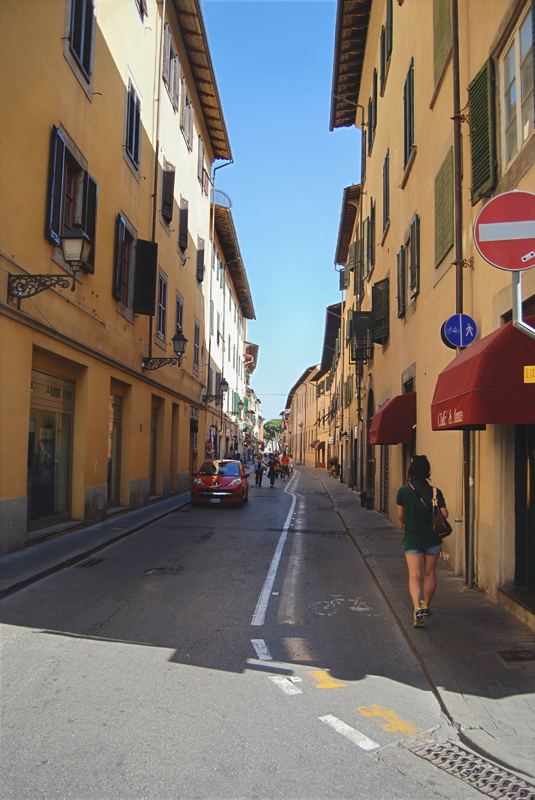 1 - A person walking down a narrow, sunlit Italian street lined with historic multi-story buildings and traditional shuttered windows